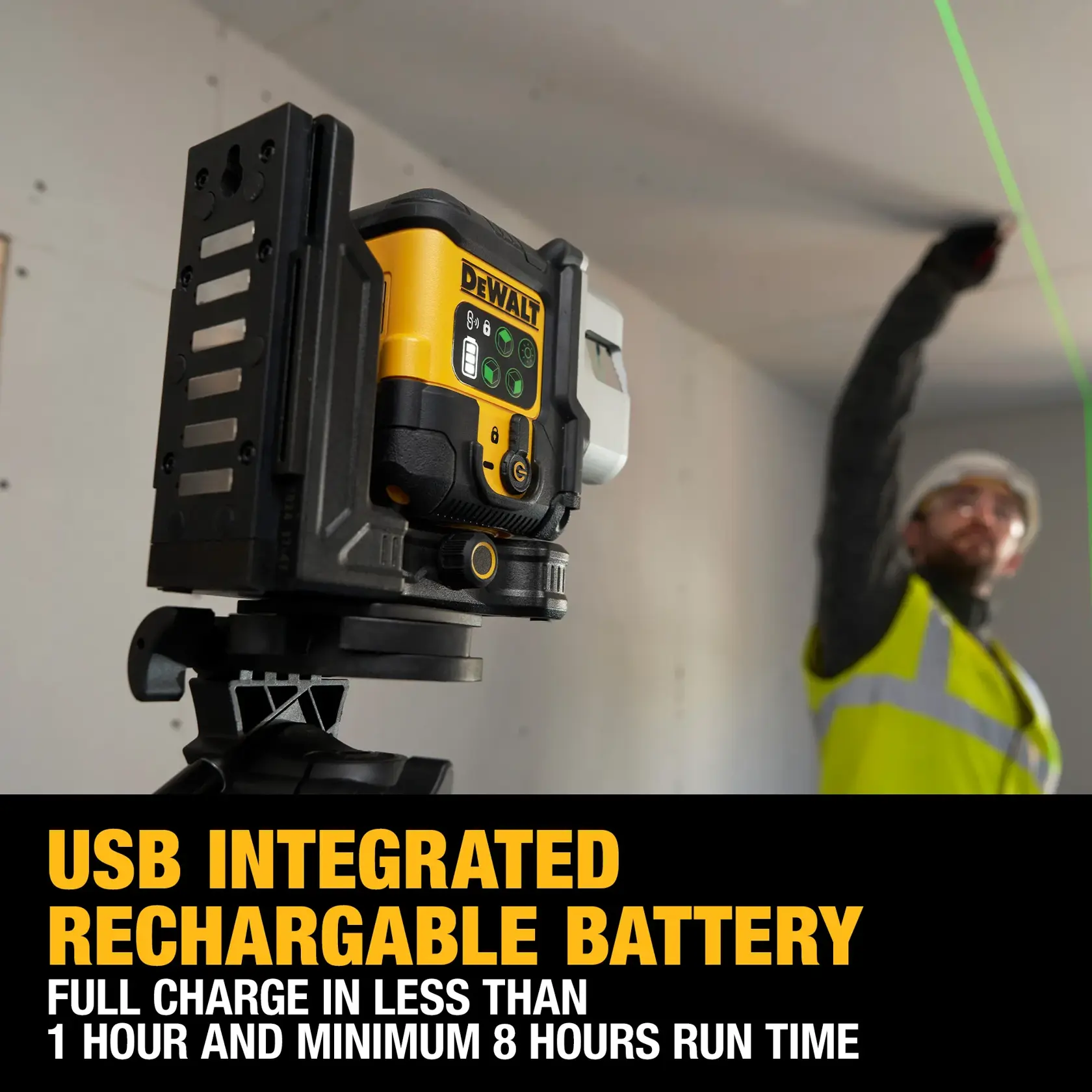 A DEWALT laser level device mounted on a tripod emits a green laser line across the room. In the background, a construction worker wearing a safety vest and helmet is installing wooden cabinets. The scene appears to be inside a partially completed room with a window and unfinished walls.