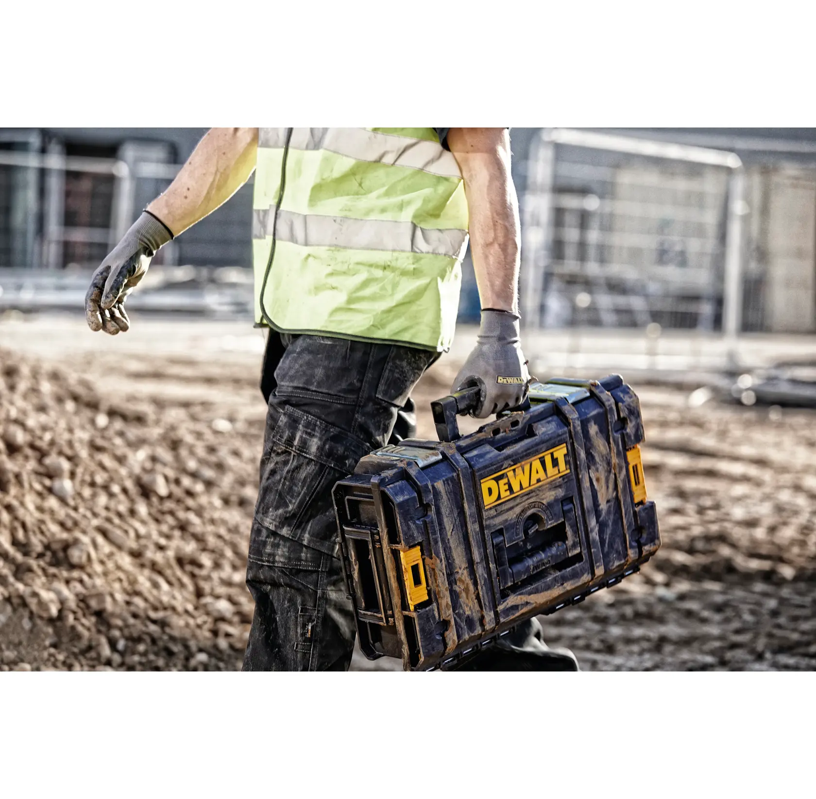 A person wearing work gloves and a high-visibility vest carries a DEWALT tool box on a construction site with dirt and fencing visible in the background.