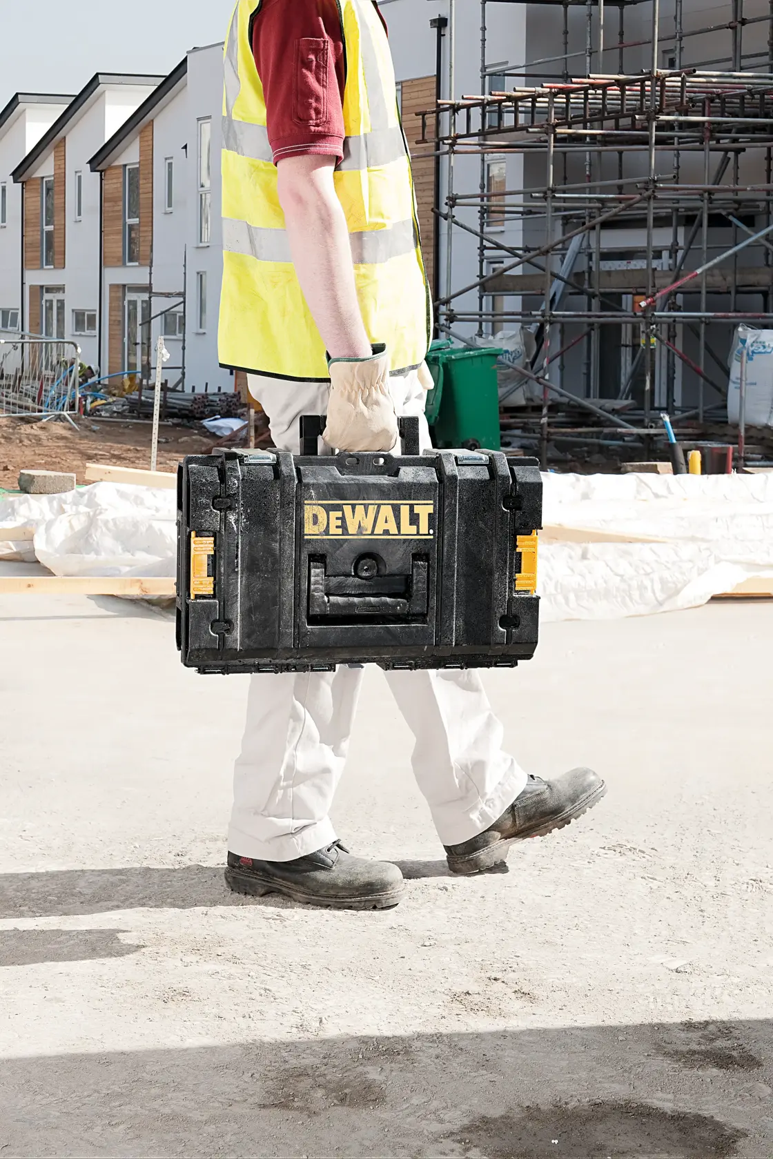 A person wearing work gloves and a yellow safety vest carries a DEWALT black tool box on a construction site, with buildings and scaffolding in the background.