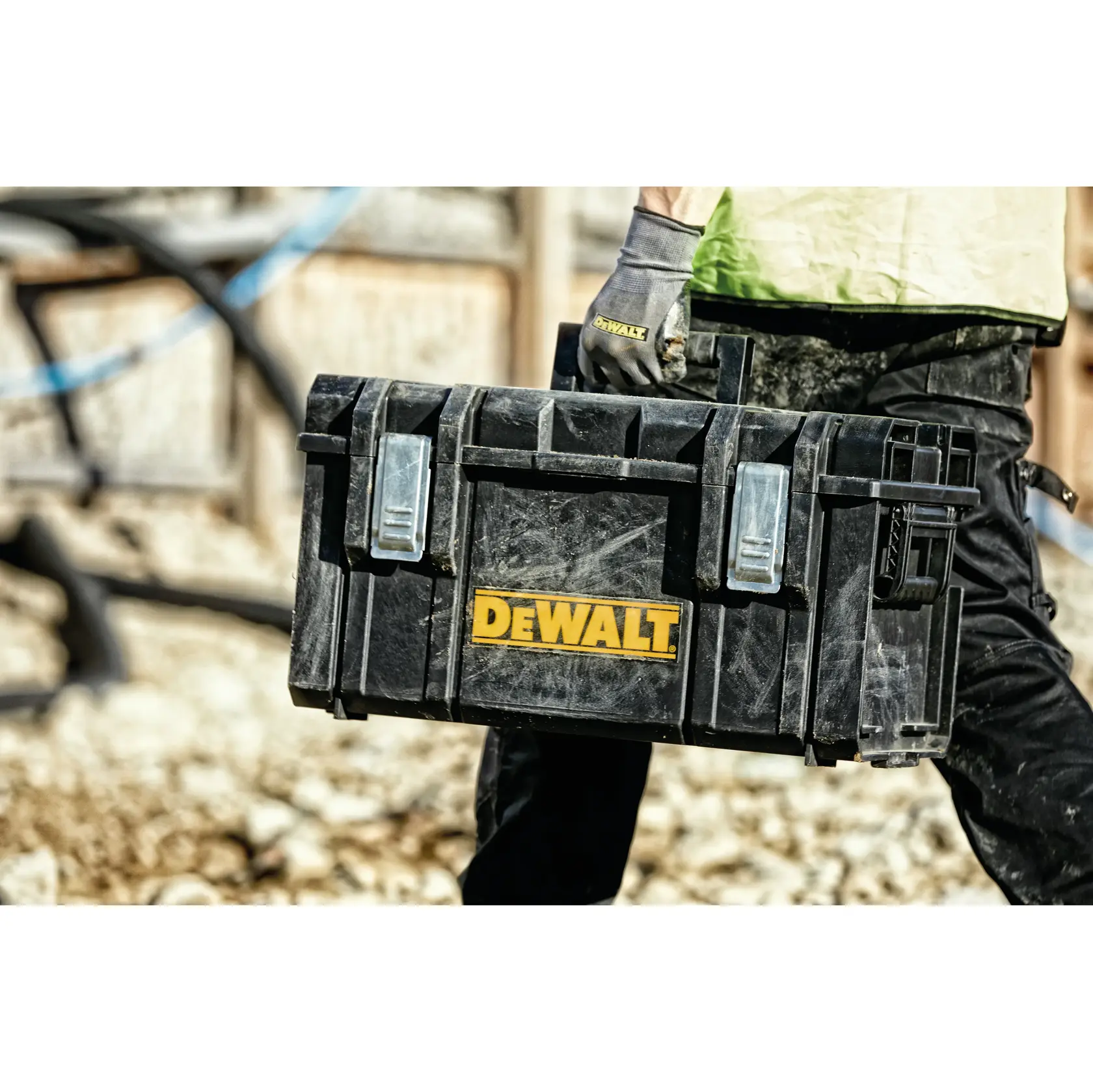 A person wearing gloves holds a DEWALT-branded black tool box with silver latches, in an outdoor construction environment.