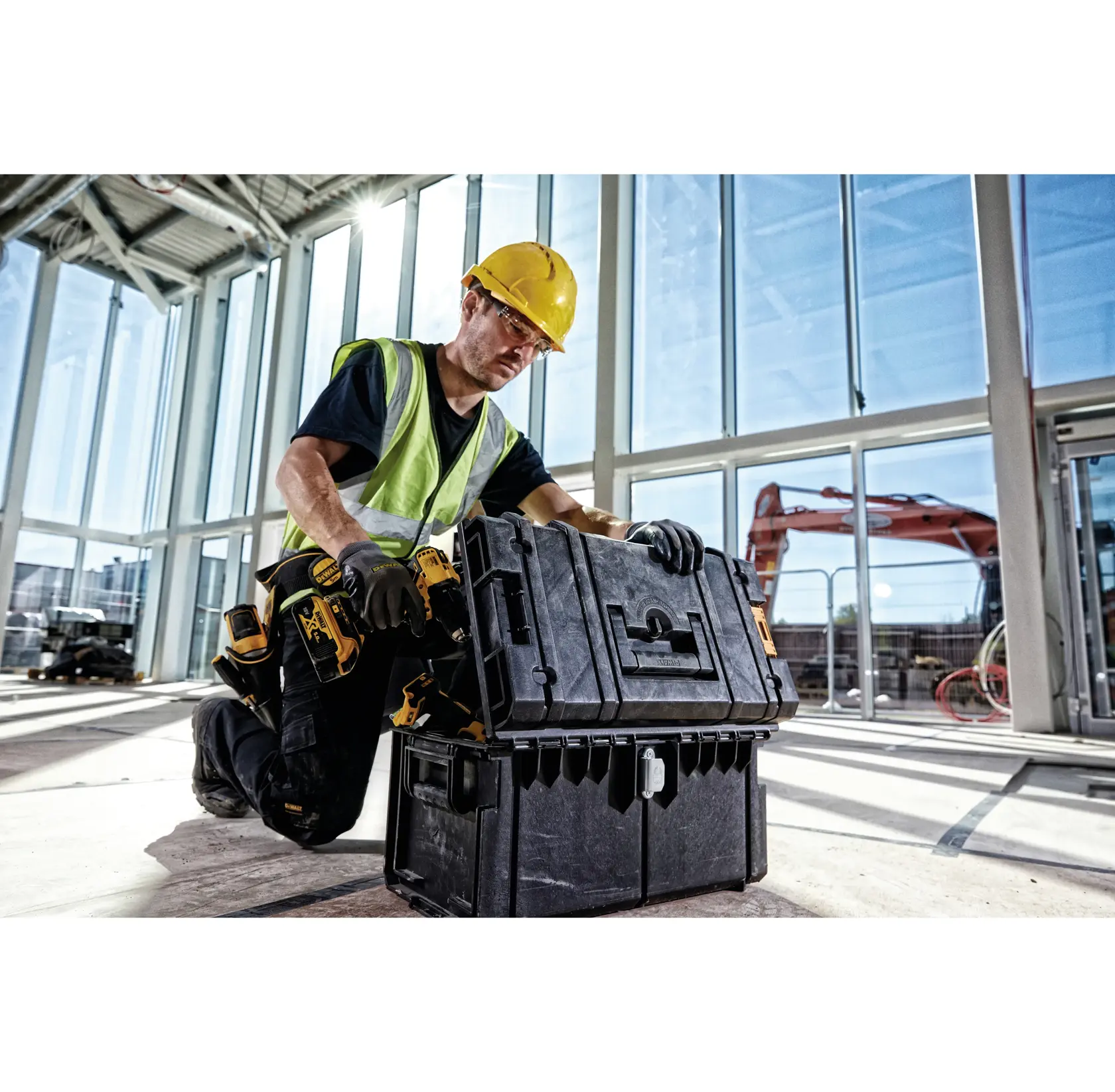 A construction worker wearing a yellow hard hat and safety vest kneels indoors on a construction site, using a DEWALT black toolbox. The worker is surrounded by tools and large windows, with construction equipment visible outside.