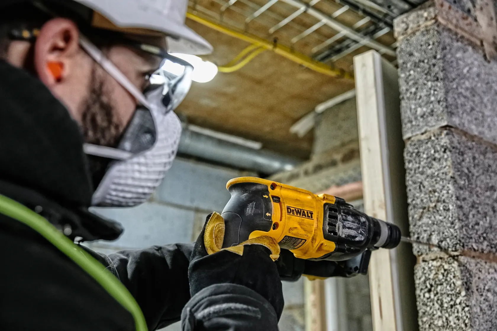 A worker wearing safety gear is using a yellow DEWALT hammer drill (model D25133K) to drill into a concrete wall at a construction site.