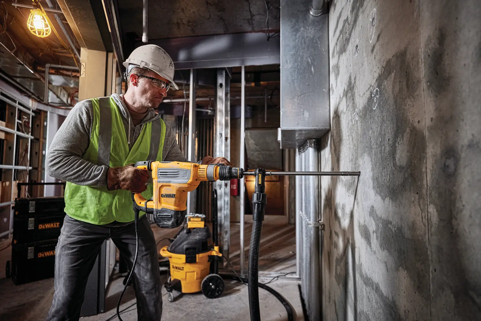 A construction worker wearing a yellow hard hat and safety vest uses a DEWALT rotary hammer drill (model D25614K) to break through a brick wall, with dust being expelled through an attached dust extraction system.