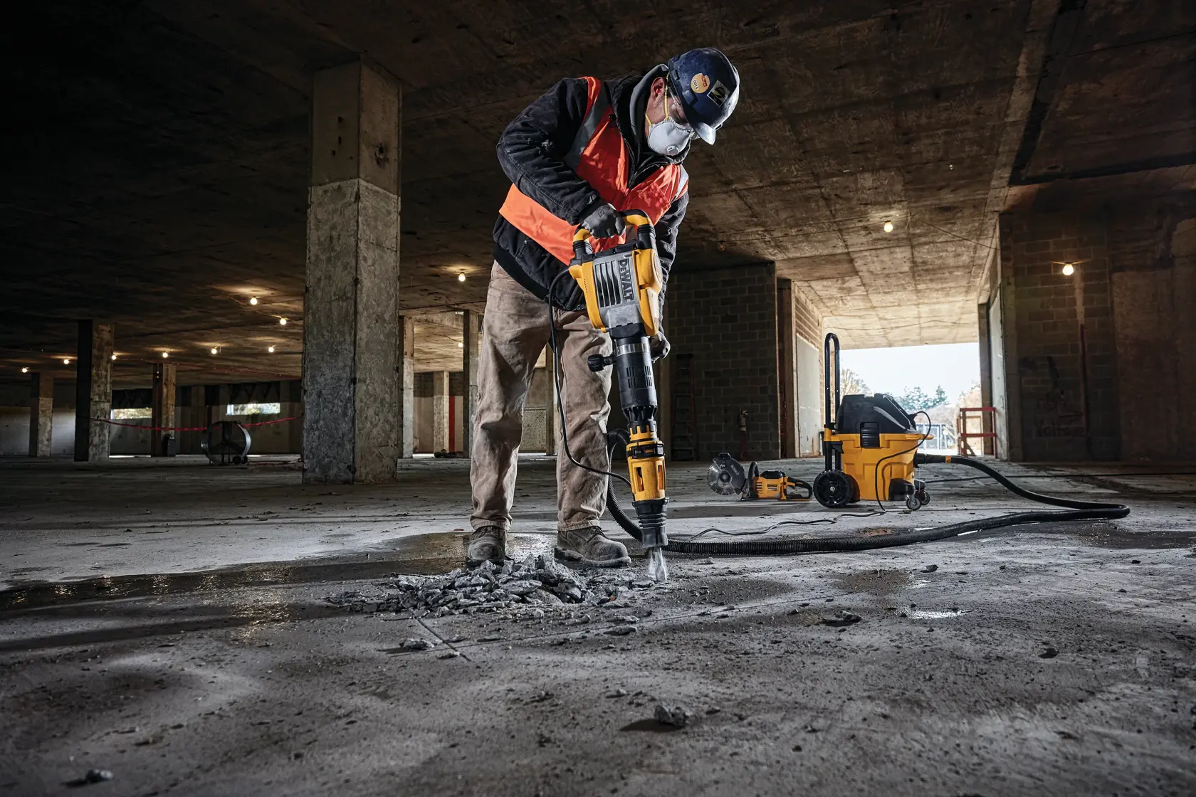 A construction worker wearing safety gear uses a DEWALT demolition hammer to break concrete in an unfinished building. A DEWALT dust extractor is visible in the background.