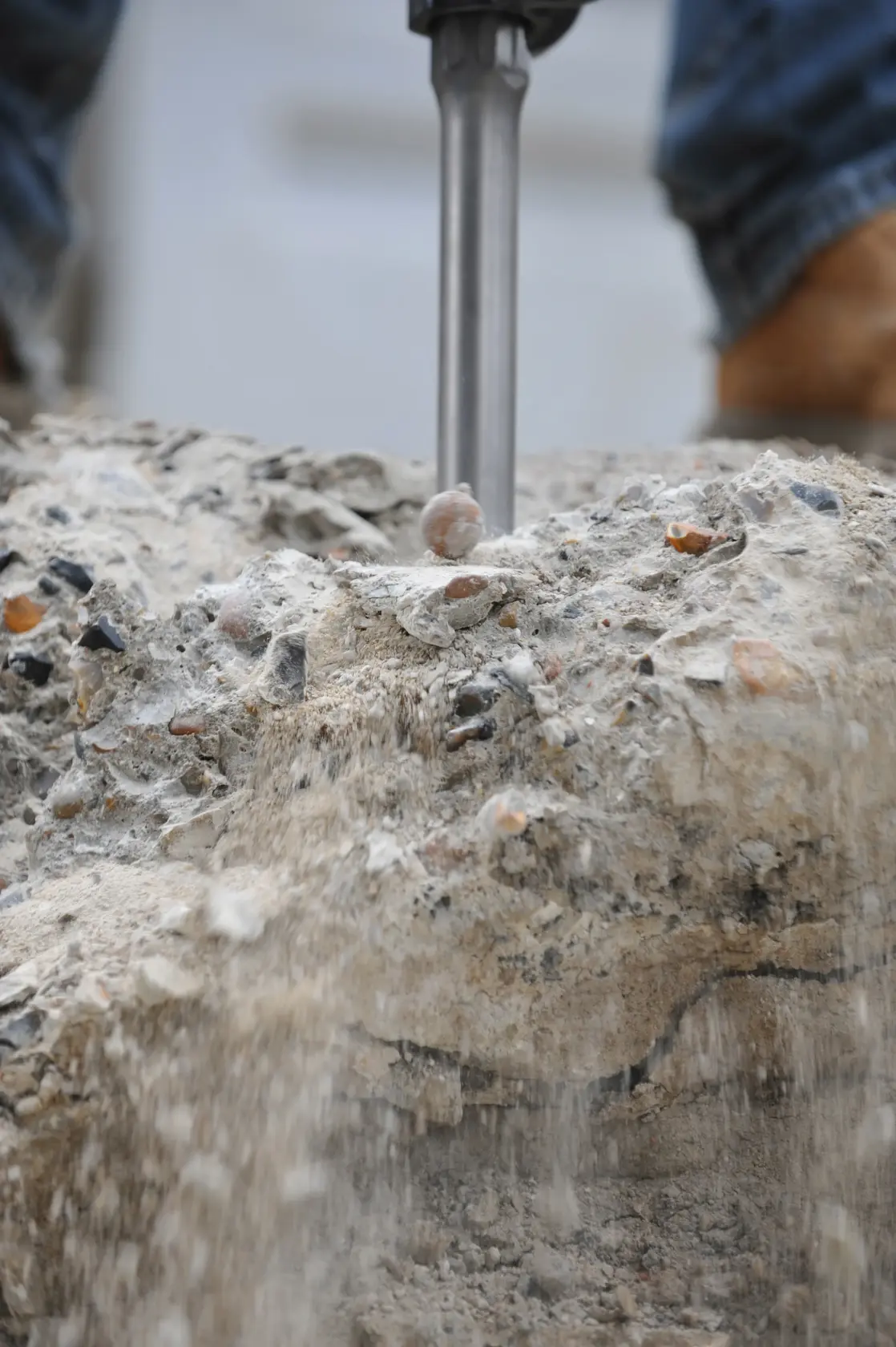 Close-up of a demolition tool breaking concrete, with dust and debris falling, and a person's feet visible in the background.