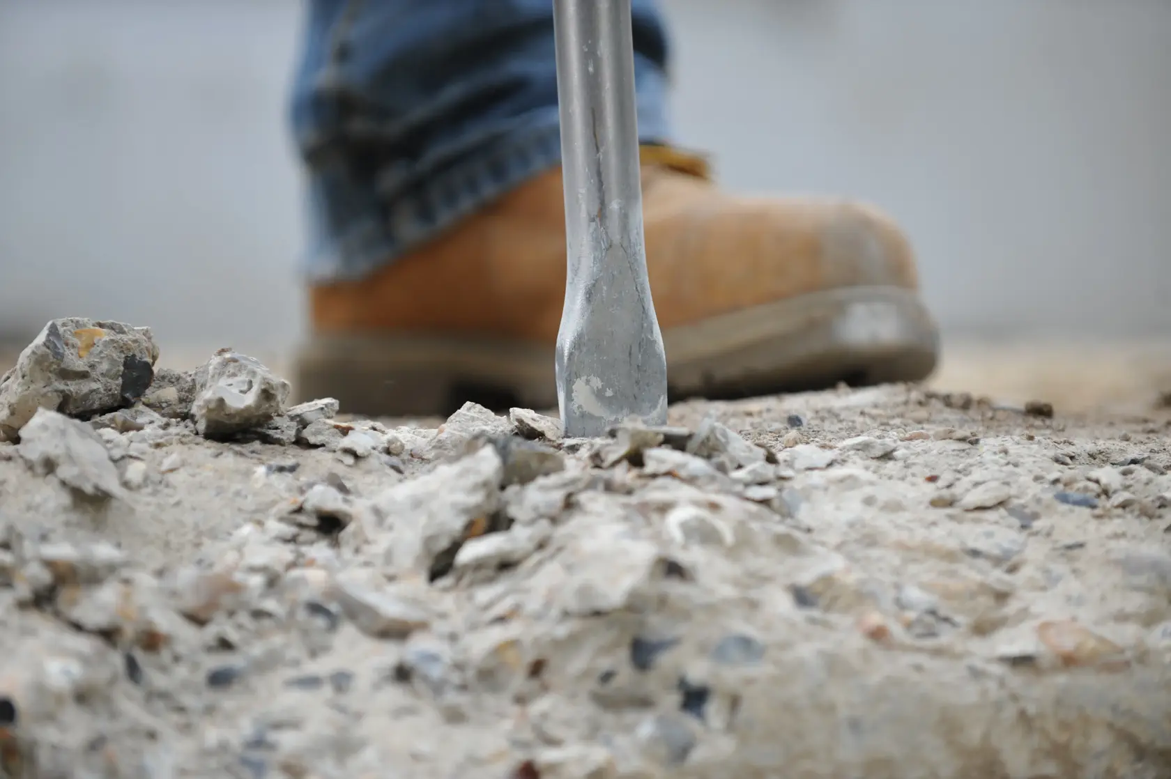 Close-up of a chisel tip breaking concrete, with a worker wearing a brown safety boot in the background.