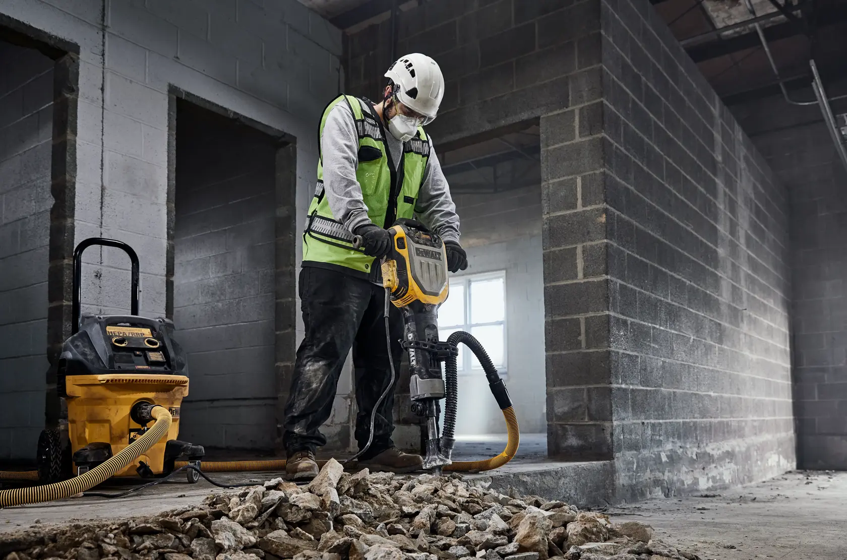 A construction worker wearing a safety vest and helmet is using a DEWALT jackhammer (model D25966) to break up concrete inside an unfinished building. There is a DEWALT dust extractor connected to the jackhammer, and broken concrete rubble is scattered on the floor.
