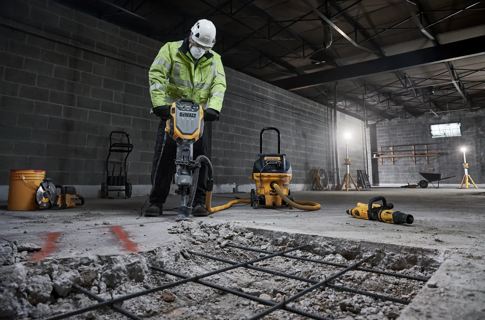 A worker wearing a safety helmet and a high-visibility jacket uses a DEWALT demolition hammer to break up concrete in an industrial setting. Various construction tools and equipment, including a vacuum and power tools, are visible in the background.