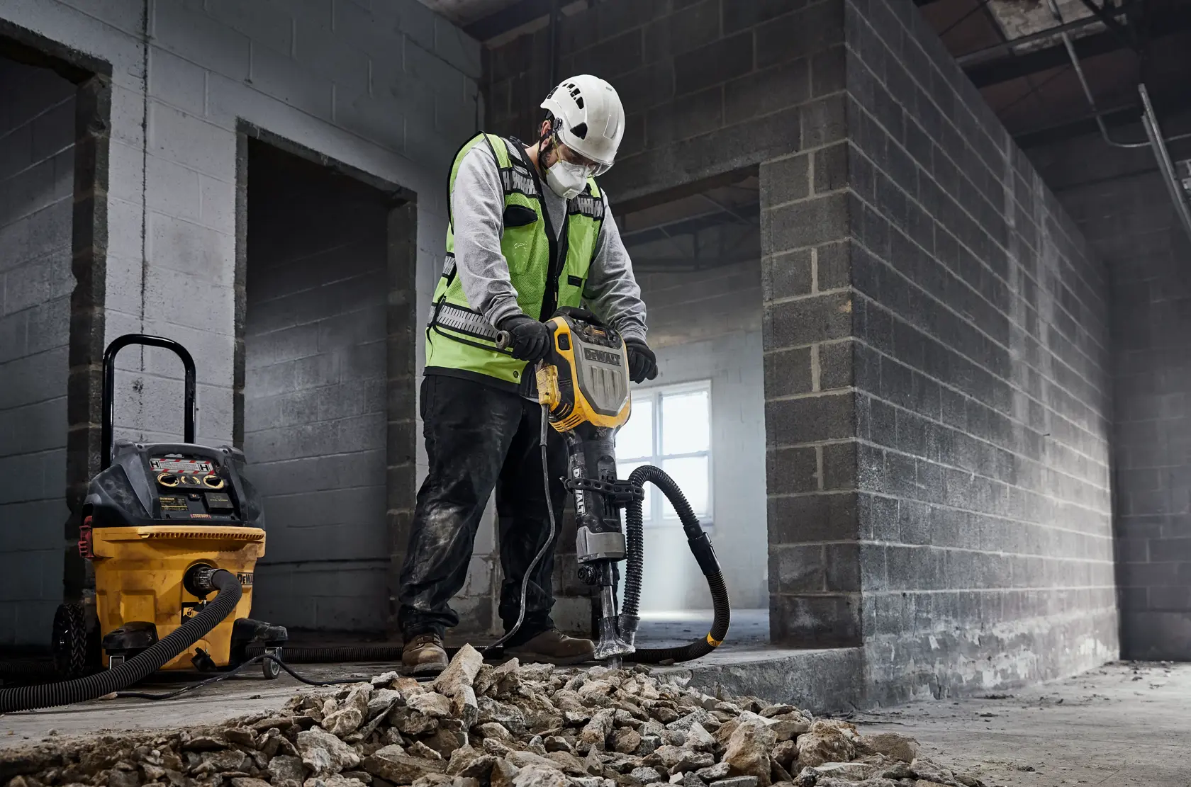 A construction worker wearing a white helmet and green safety vest is operating a DEWALT jackhammer to break up concrete and rocks inside an unfinished building. Nearby, a yellow DEWALT vacuum is connected to the jackhammer for dust extraction.