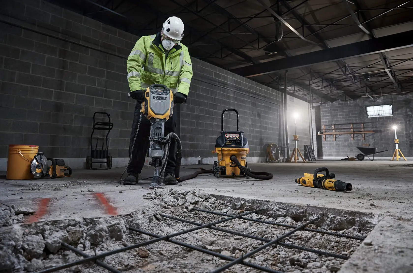A construction worker using a DEWALT D25966 demolition hammer to break concrete in an industrial indoor setting. The worker is wearing a safety helmet and high-visibility jacket. Additional DEWALT tools and equipment, such as a vacuum, drills, and a bucket are visible in the background.