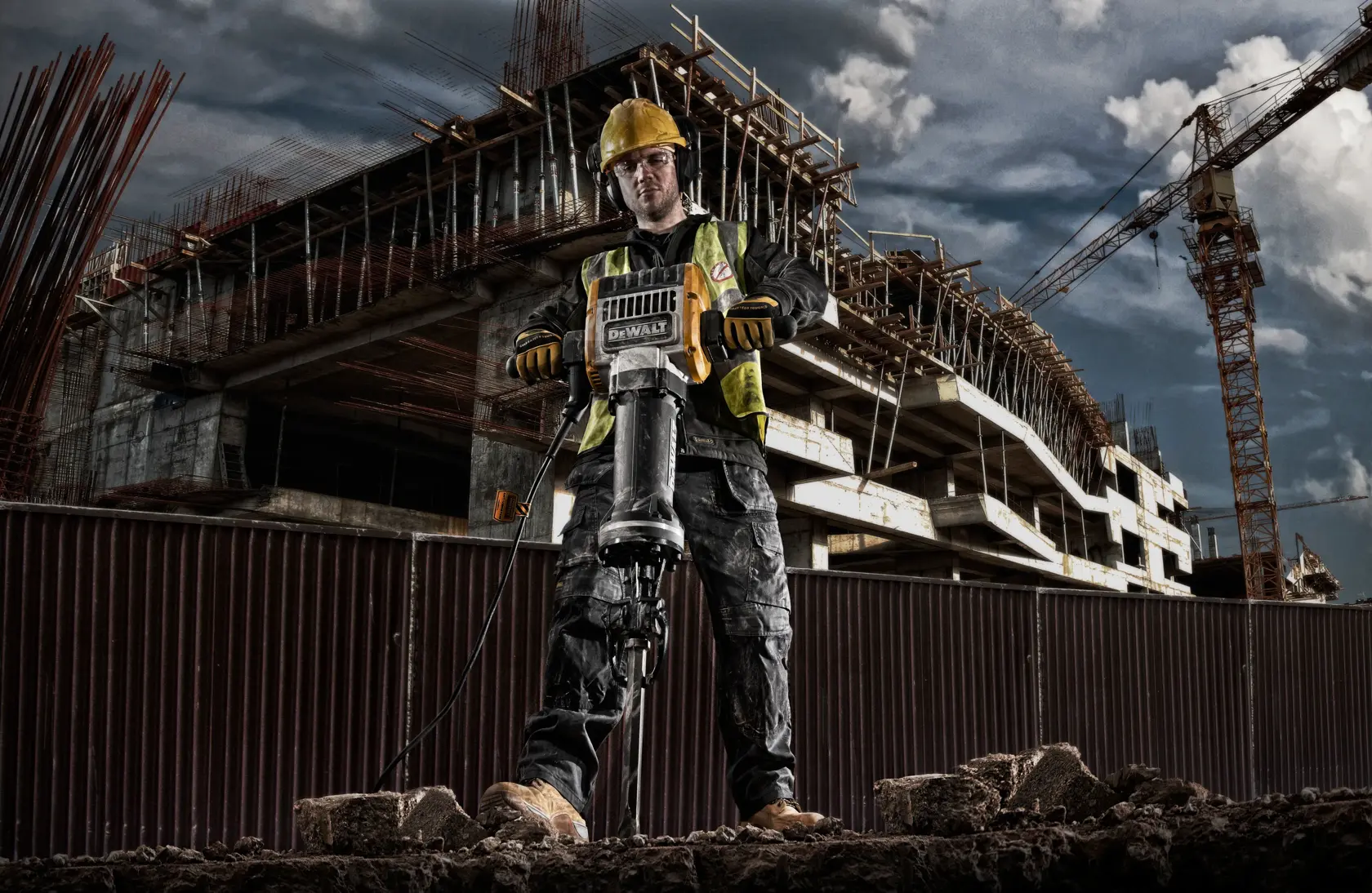 A construction worker using a DEWALT D25981 demolition hammer on a construction site, with a partially built structure and cranes in the background.