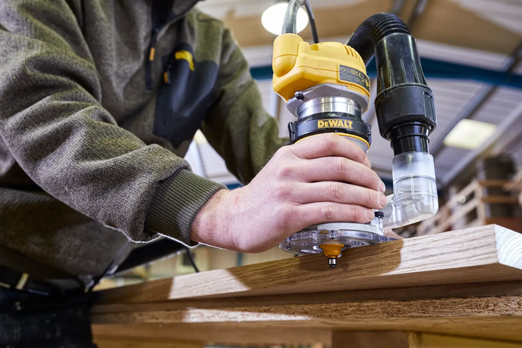 A person is using a DEWALT router tool, model D26200, to shape wood in a workshop. The router has a dust extraction attachment connected.