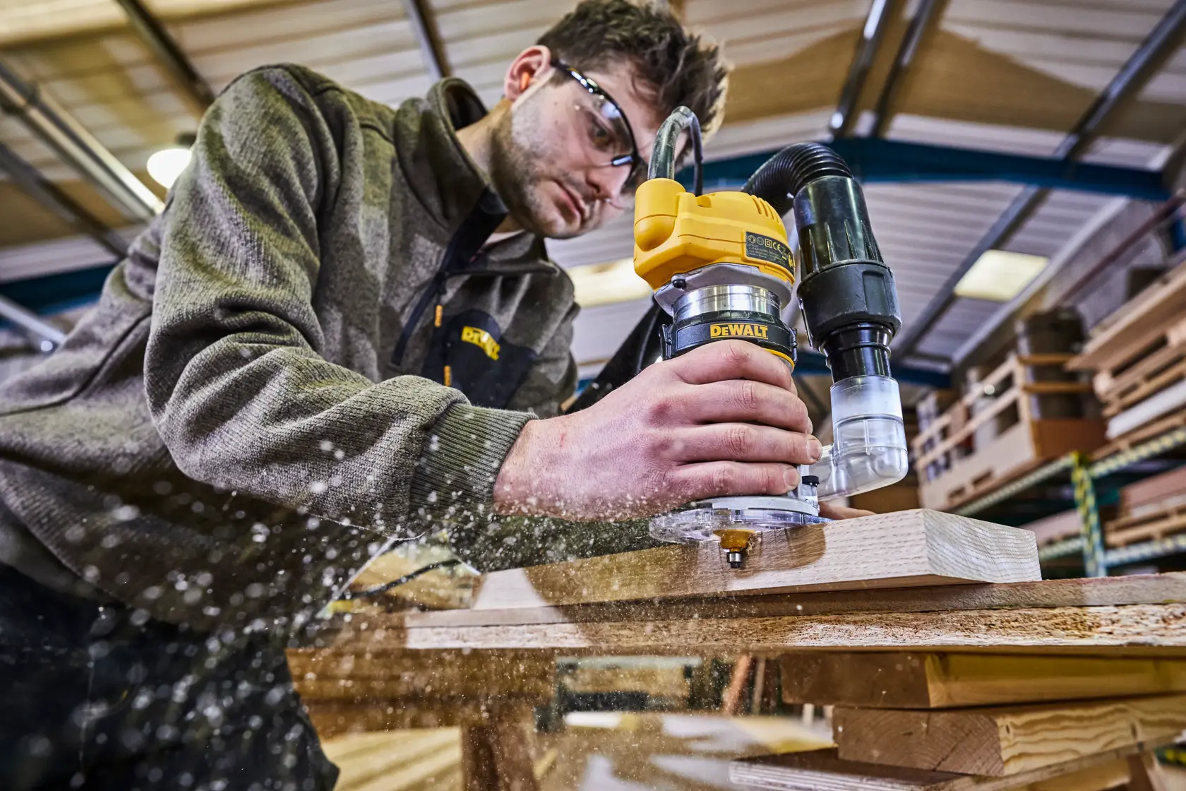 A person using a DEWALT D26200 router on a piece of wood, with sawdust flying, in a woodworking workshop.
