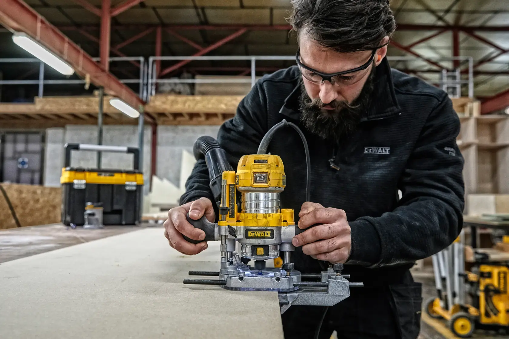 A person using a yellow DEWALT D26204K router tool on a piece of wood in a workshop. The individual's face is obscured, and there are various woodworking equipment and storage boxes in the background.