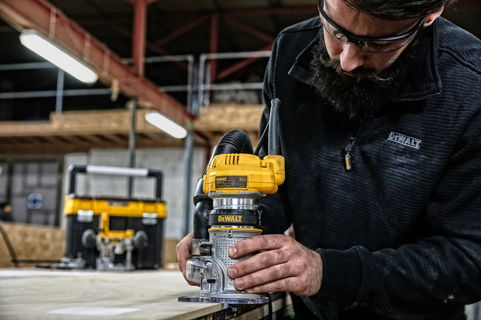 A person using a DEWALT D26204K router tool on a wooden surface in a workshop, with the person's face blurred for privacy. There is another DEWALT tool visible in the background.