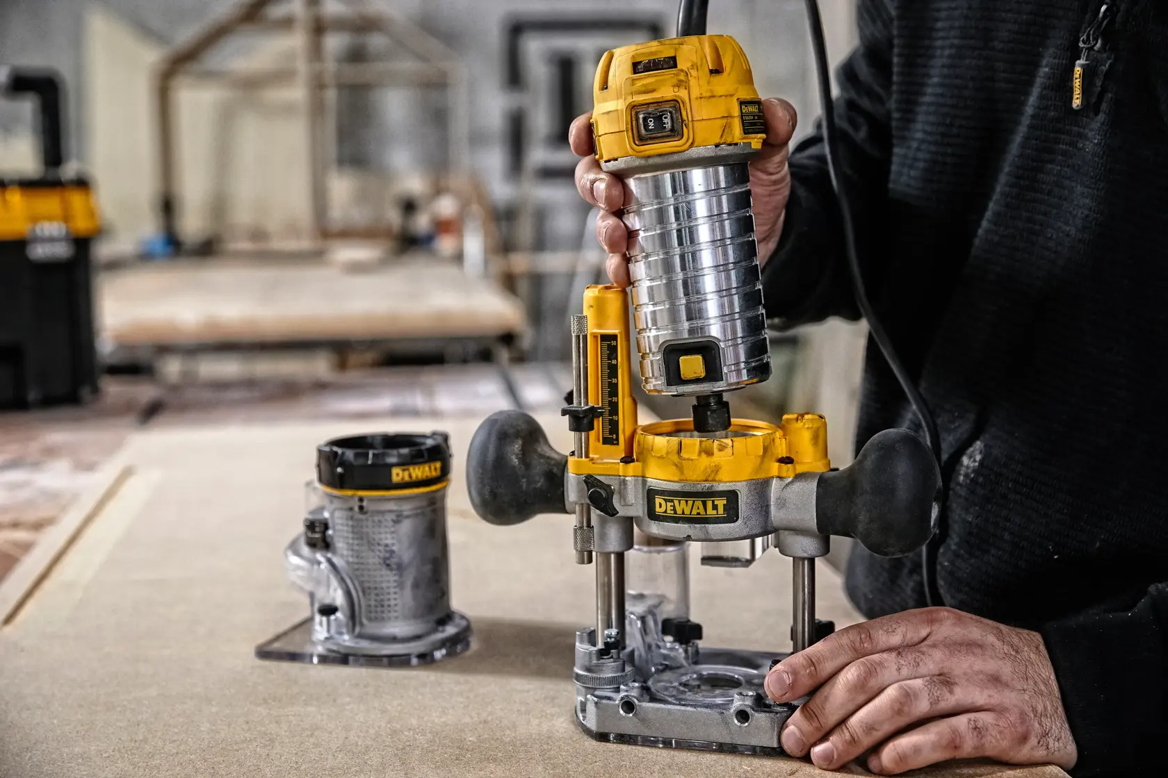 A person is using a DEWALT D26204K router tool on a piece of wood in a workshop. Another base attachment is visible on the table. The focus is on the yellow and silver DEWALT router being operated.