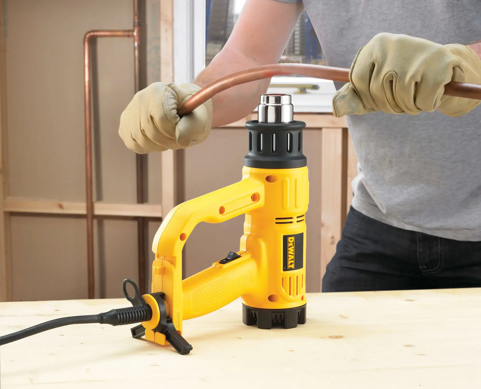 A person wearing gloves uses a DEWALT heat gun to bend a copper pipe on a wooden work surface.