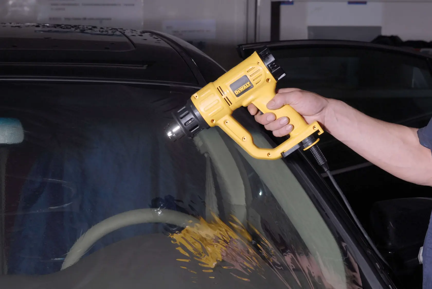 A person using a DEWALT heat gun (model D26411) to apply heat on the windshield of a car.