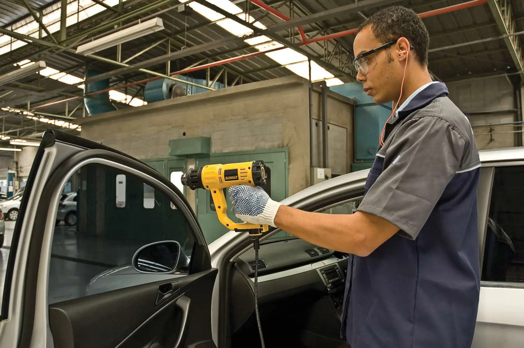 A person wearing gloves is using a yellow DEWALT heat gun (model D26411) on the interior of a car door in a workshop setting. The face of the person is blurred for privacy.