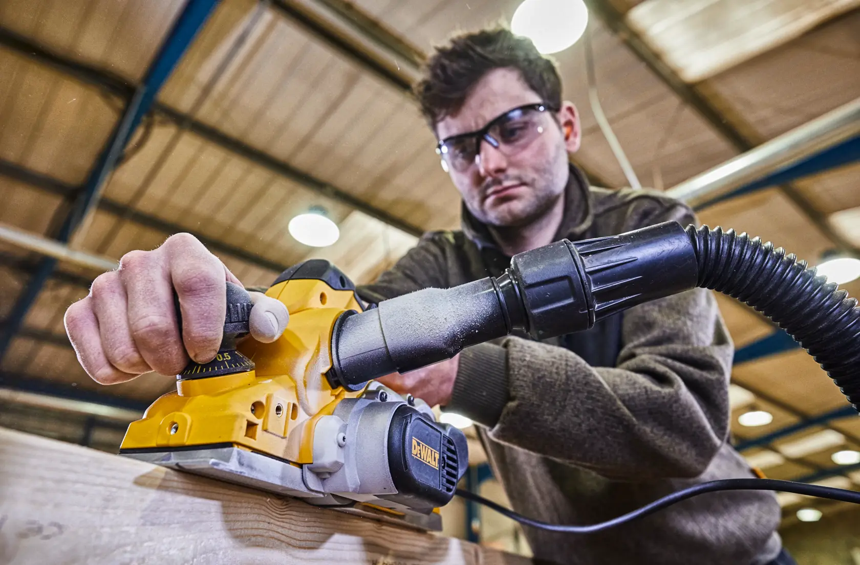A person using a DEWALT electric planer (model D26500K) on a piece of wood in a workshop, with a dust extraction hose attached. The person's face is blurred.