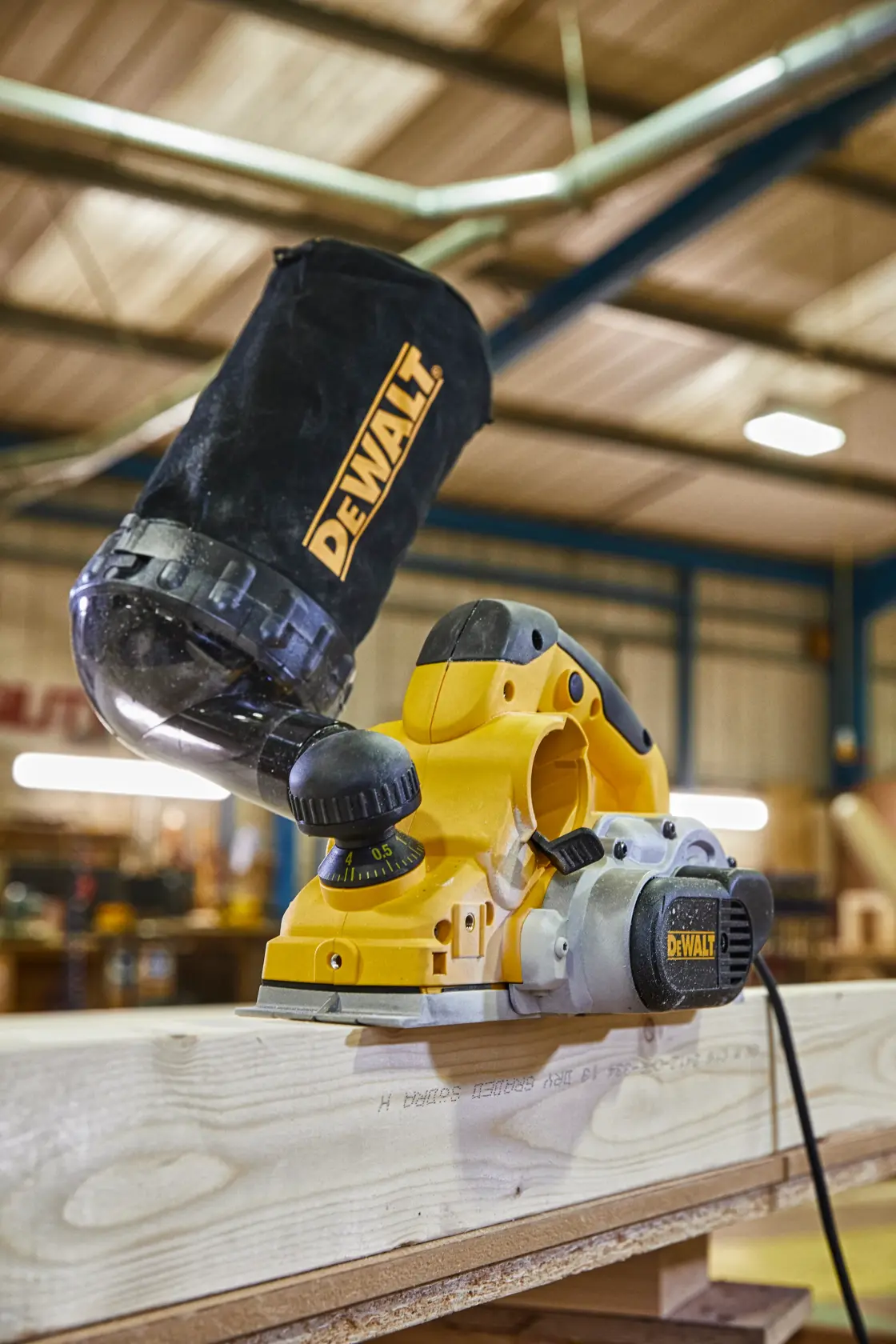 A close-up of a DEWALT electric hand planer being used on a wooden plank in a workshop. The planer has a dust collection bag attached, and the workspace background is blurred.