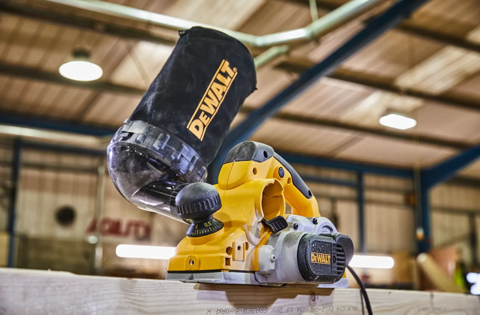 A DEWALT electric planer with a dust collection bag attached, shown in use on a wooden plank in a workshop setting.