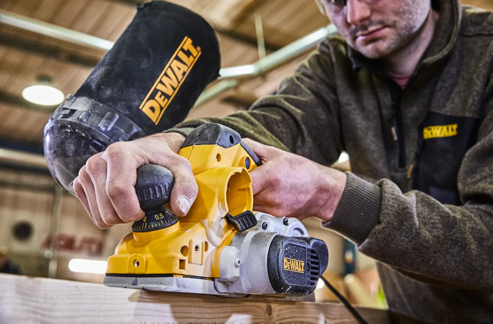 A person using a DEWALT electric planer, model D26500K, to smooth a piece of wood. The planer is yellow and black, with DEWALT branding visible, and the scene is set in a workshop environment. The person's face is blurred for privacy.