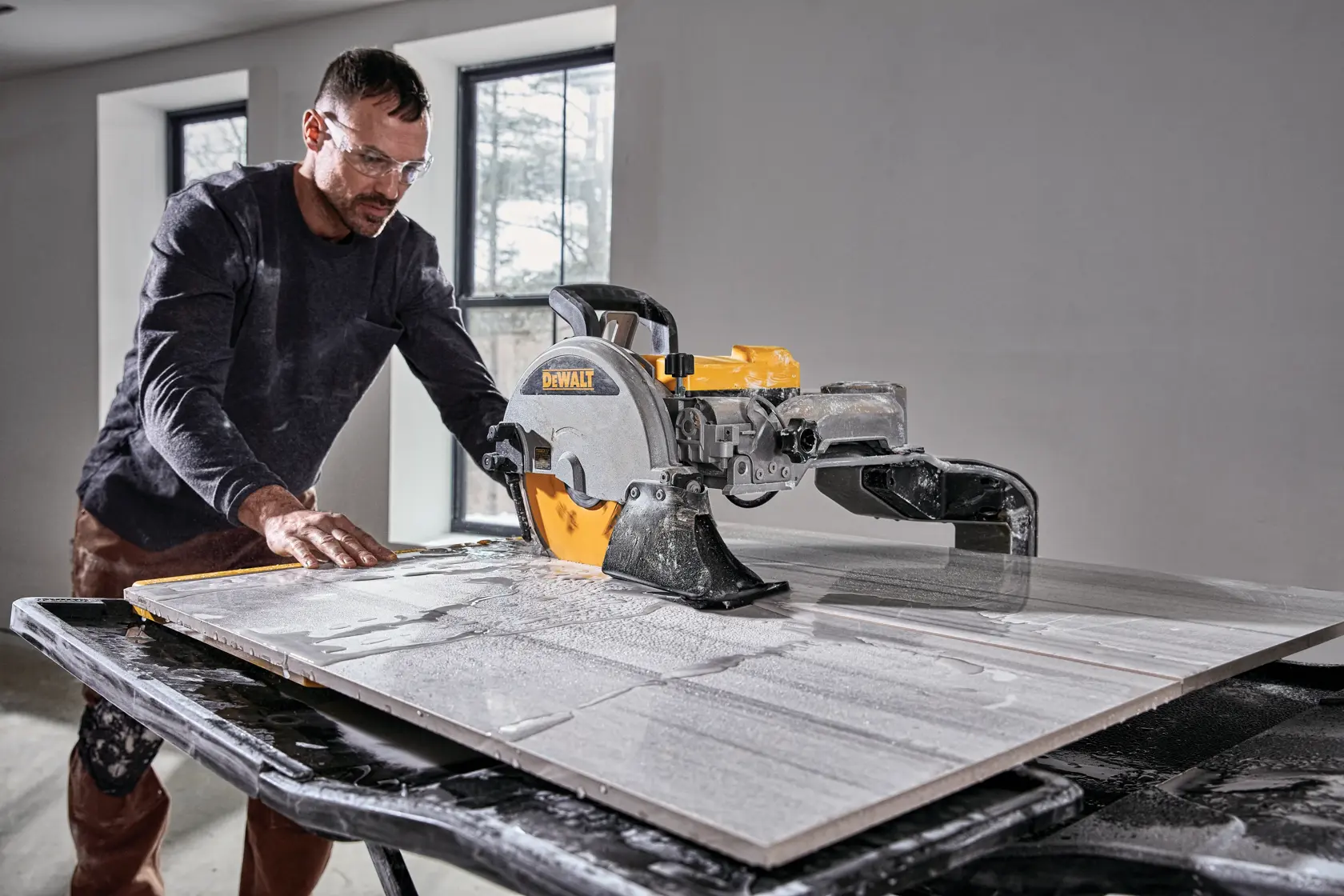 A person using a DEWALT D36000 tile saw to cut a large tile on a worktable in a room with two windows.