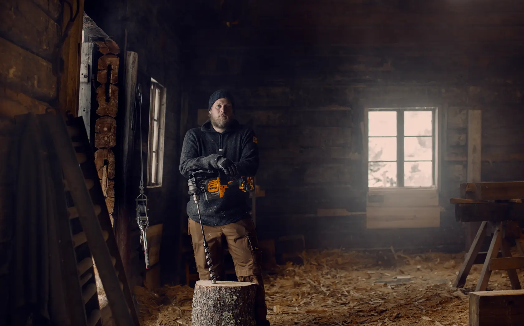 A person standing in a rustic workshop holding a yellow DEWALT DCD460N power drill with a large auger bit, next to a tree stump. The person's face is blurred, and wood shavings cover the floor. Sunlight streams through a window in the background.