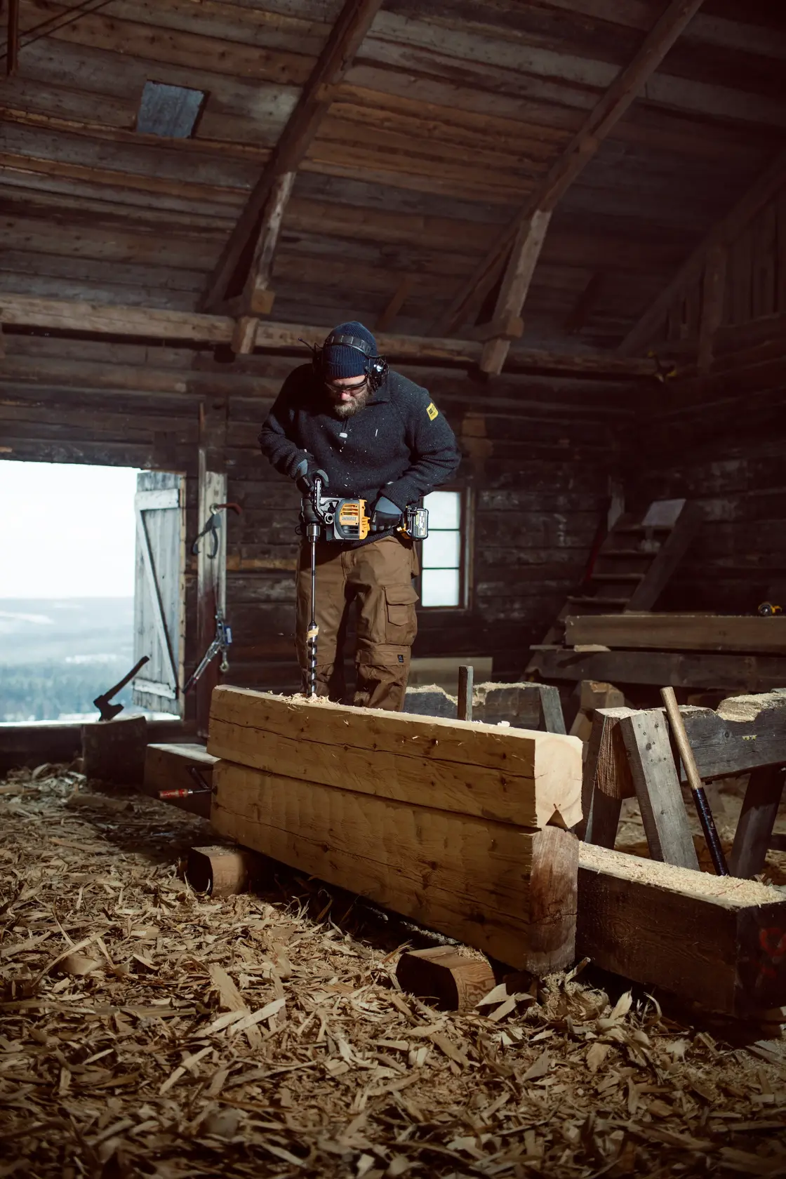 A person using a DEWALT DCD460N power drill to bore holes into a large wooden beam inside a rustic workshop. Wood shavings are scattered on the floor and several pieces of timber are visible around the workspace.
