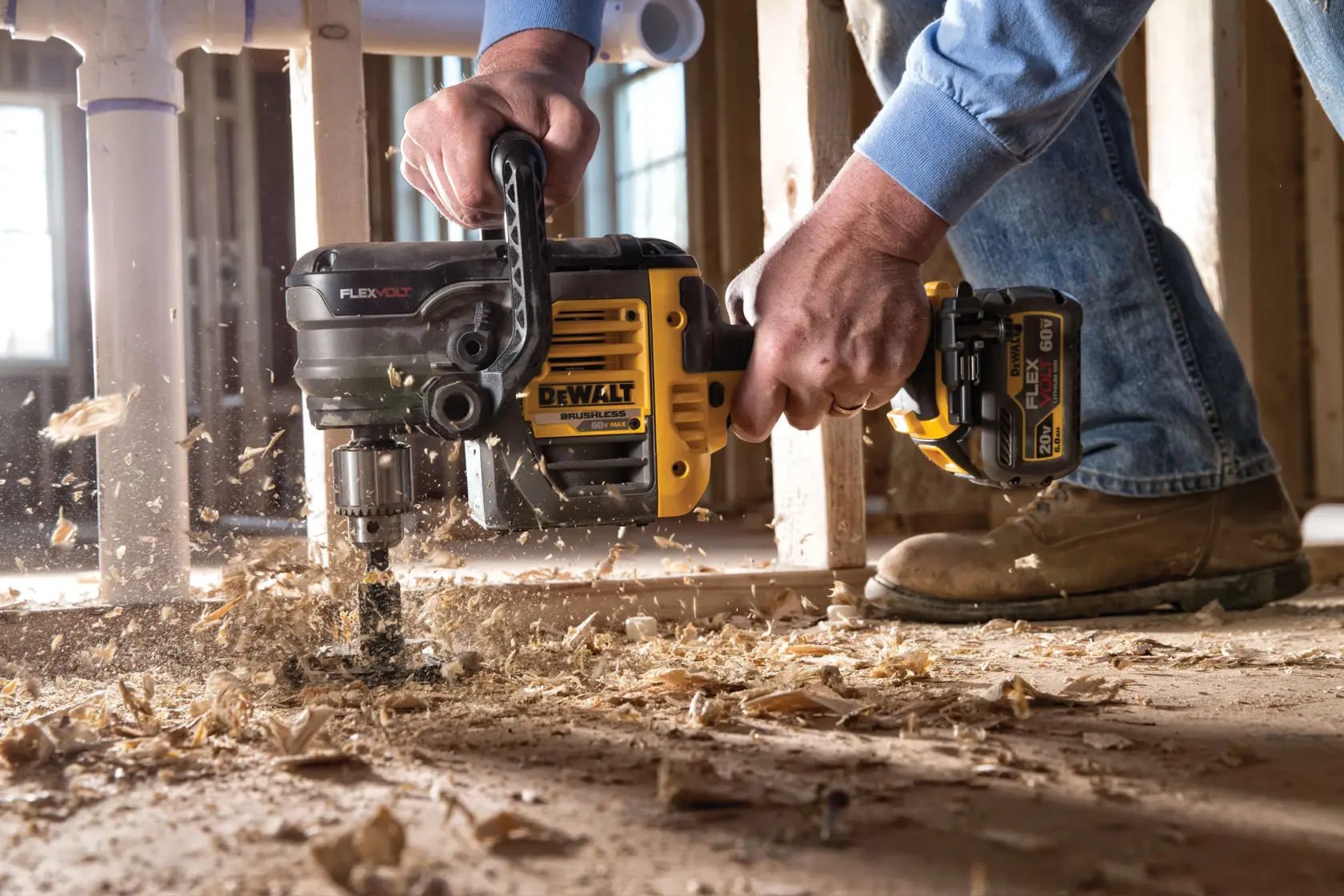 A DEWALT cordless power drill being used to bore a hole into a wooden floor, with wood shavings scattered around. The drill is being operated by a person wearing work boots and jeans, inside a construction site.