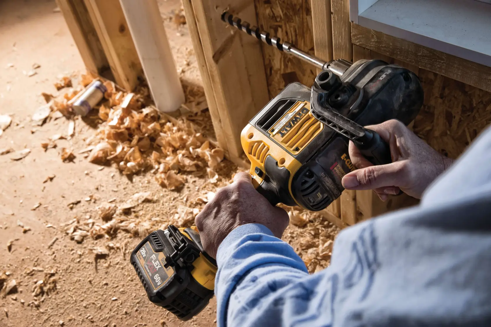 A person using a DEWALT cordless power drill to bore a hole in a wooden wall frame, with wood shavings scattered on the floor.