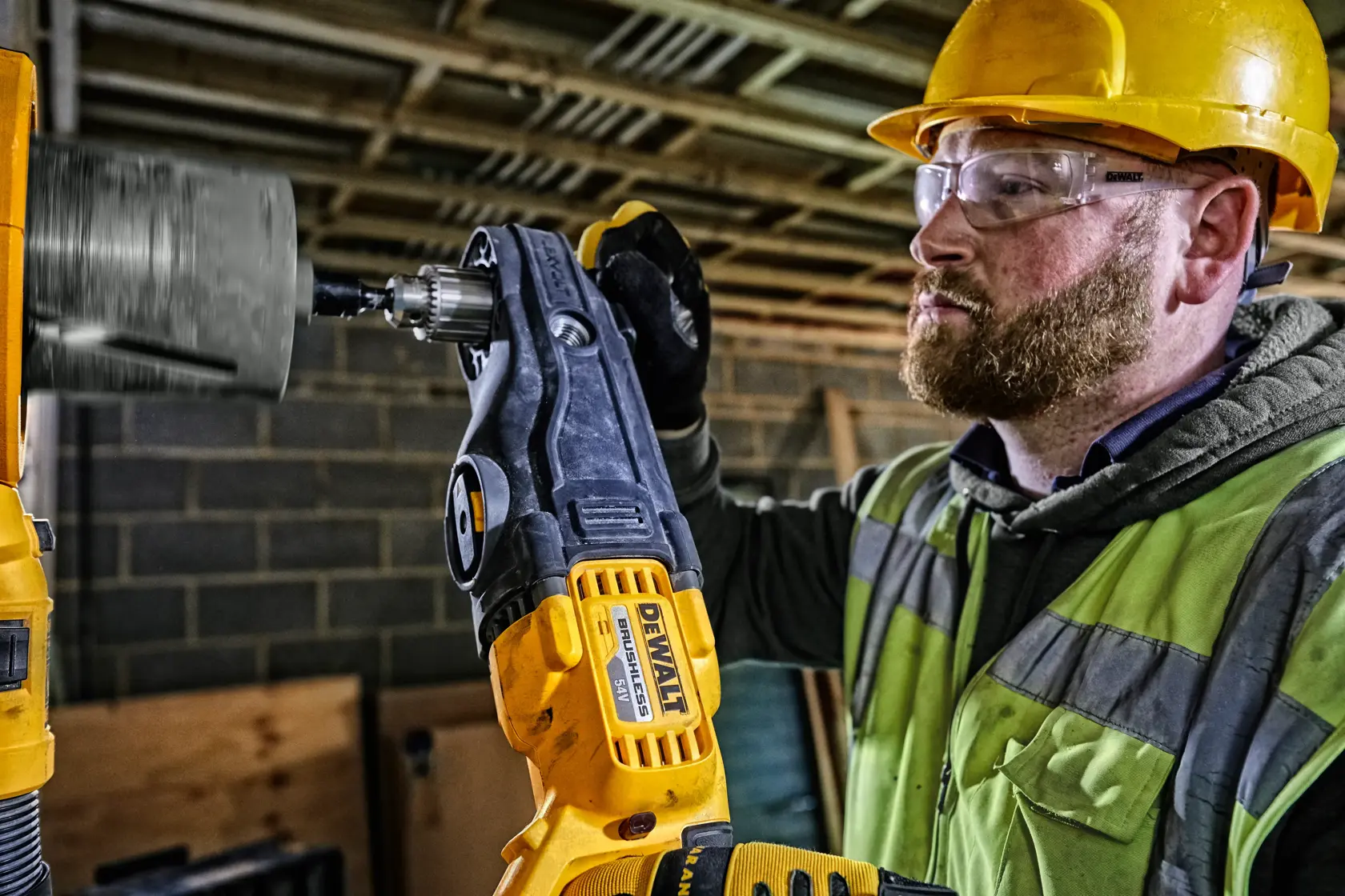 Eine Person mit gelbem Schutzhelm und Sicherheitsweste benutzt eine DEWALT DCD470N Bohrmaschine mit einem Aufsatz zum Bohren in eine Oberfläche auf einer Baustelle. Das Gesicht der Person ist aus Datenschutzgründen unscharf.