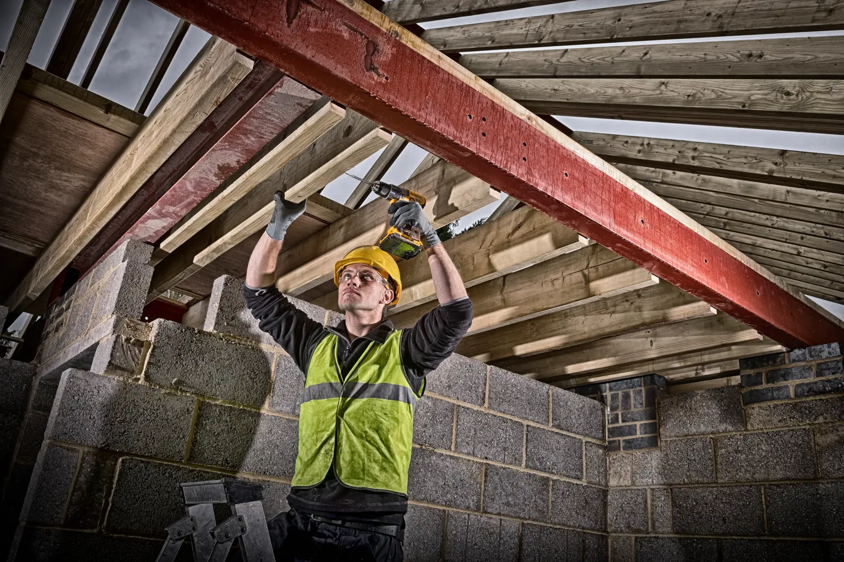 A construction worker wearing a high-visibility vest and hard hat uses a DEWALT cordless drill to secure a wooden beam in the ceiling structure of a building under construction. The worker stands on a ladder surrounded by concrete block walls and timber roof trusses.