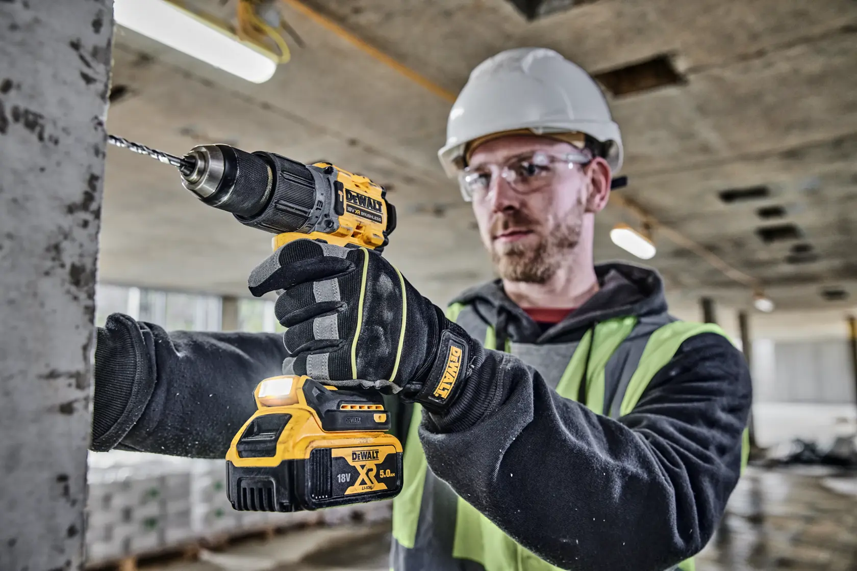 A person wearing gloves, a safety vest, and a hard hat is using a DEWALT DCD805P2T cordless drill to drill into a concrete surface at a construction site. The face is blurred for privacy.