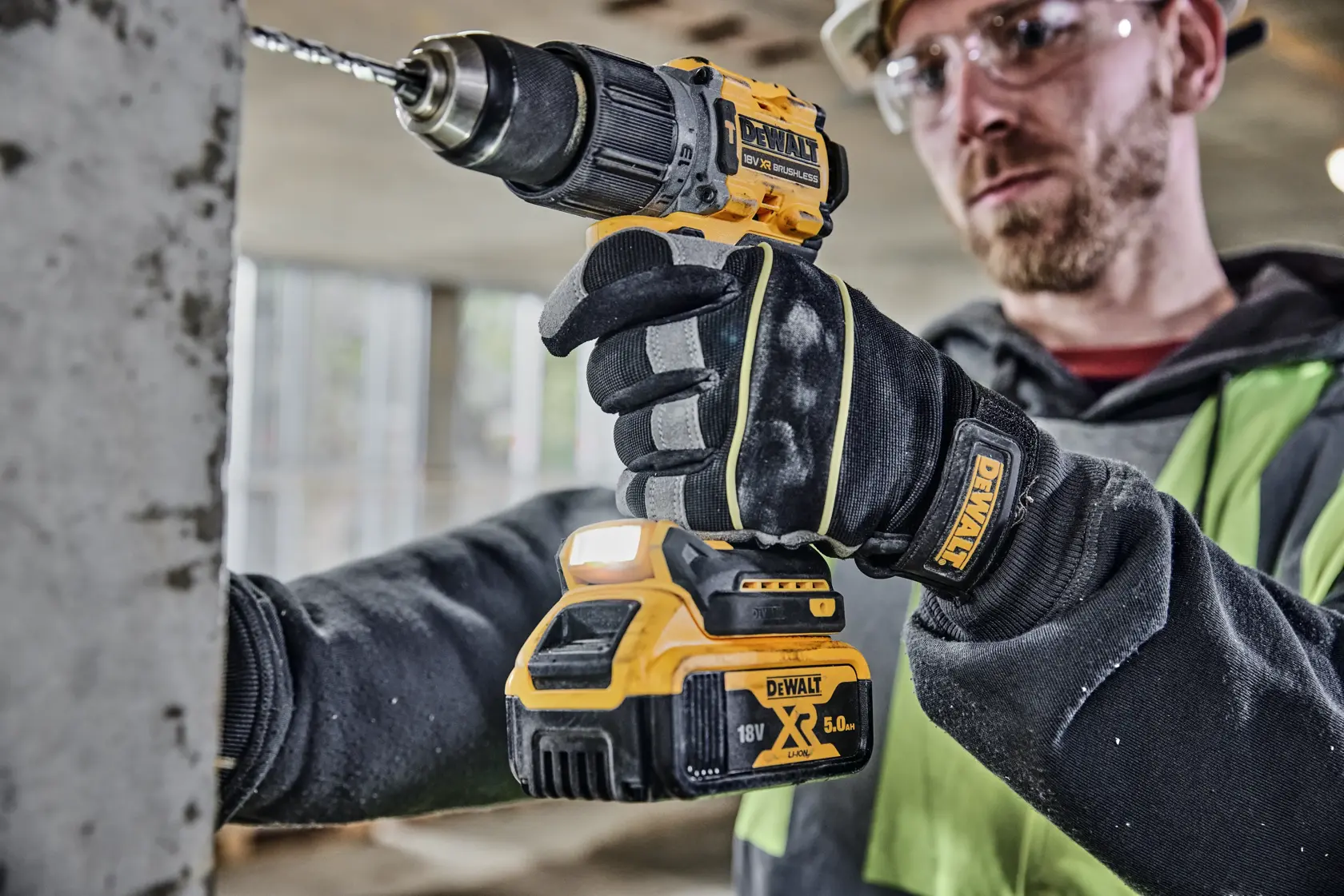 A person wearing DEWALT-branded gloves is using a DEWALT 18V XR cordless drill driver (model DCD805P2T) to drill into a concrete surface in an indoor construction setting. The face of the person is blurred.