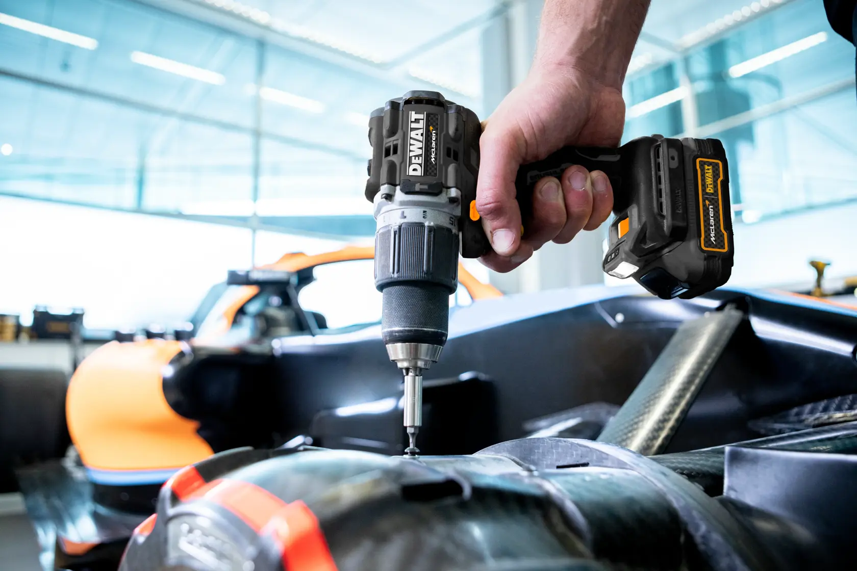 A close-up of a DEWALT cordless drill being used by a person to work on a part of a vehicle, with an orange car visible in the background in a bright, modern workshop.