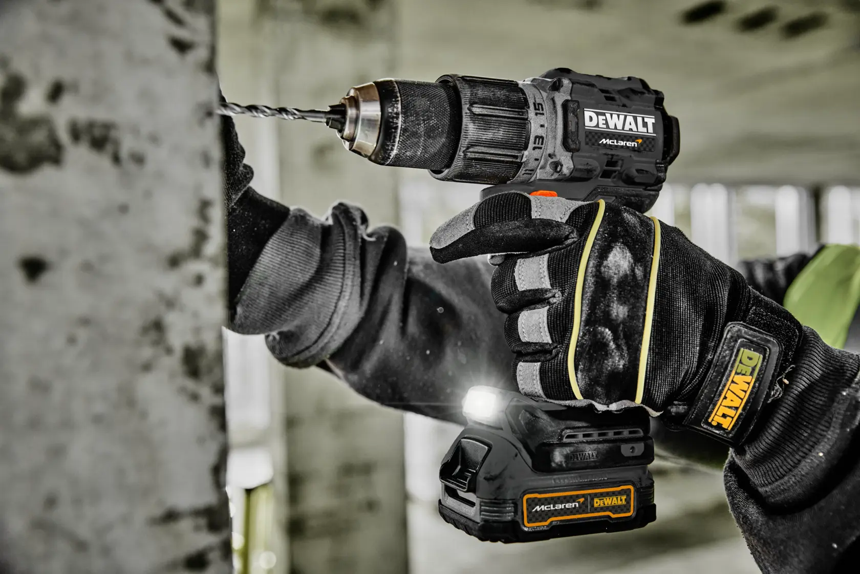 A person wearing protective gloves is using a DEWALT cordless drill to drill into a concrete wall. The drill has a visible LED light and DEWALT branding. The scene takes place in an unfinished building or construction site.