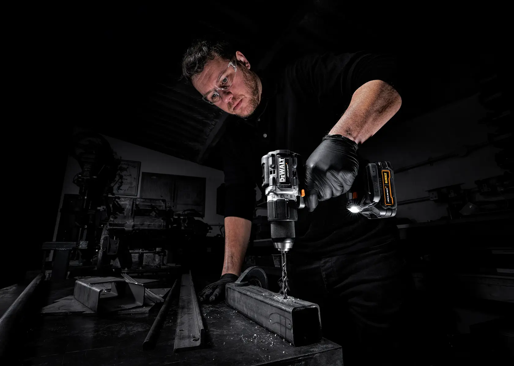 A person wearing black gloves is using a DEWALT cordless drill to drill into a metal beam in a workshop setting. The focus is on the power tool and the drilling process, with metal shavings visible on the surface.