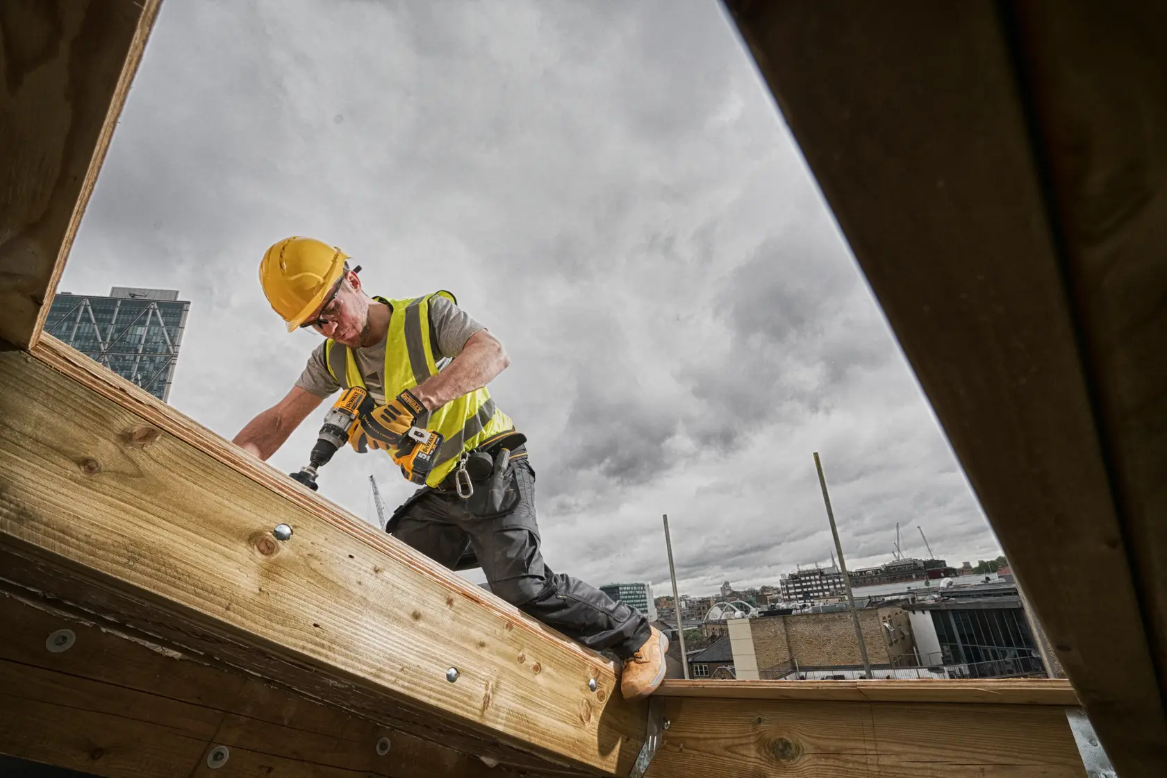 A construction worker wearing a yellow hard hat and high-visibility vest uses a DEWALT cordless drill while working on a wooden beam outdoors, with buildings visible in the background under a cloudy sky.