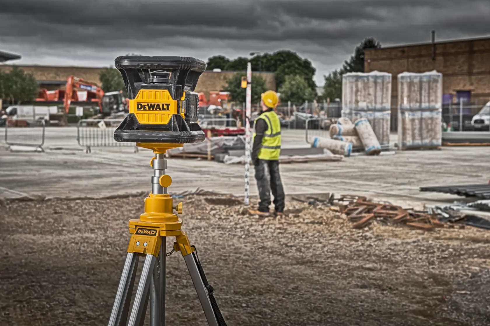 DEWALT laser level on tripod at a construction site with a worker in safety gear and equipment in background.