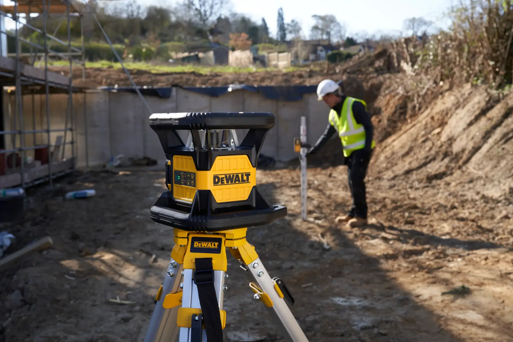 DEWALT laser level on tripod at construction site, worker in safety gear measuring ground in background.