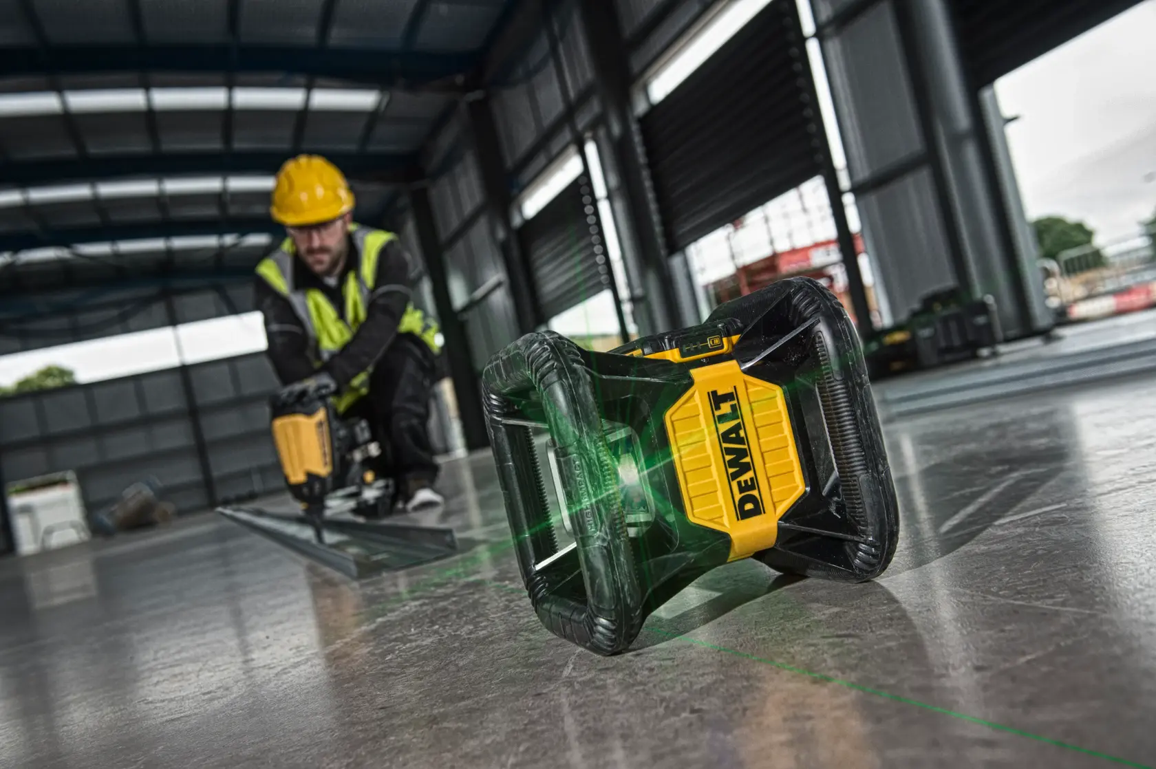 DEWALT laser level projects a green line on a construction site floor, worker in background with power tool.