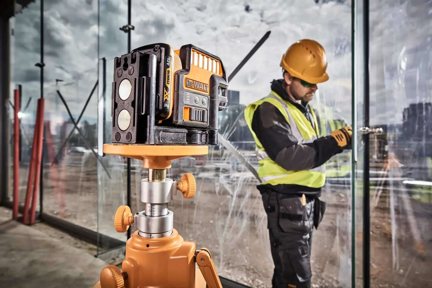 DEWALT green laser level on tripod at construction site, worker installing glass panel in background.