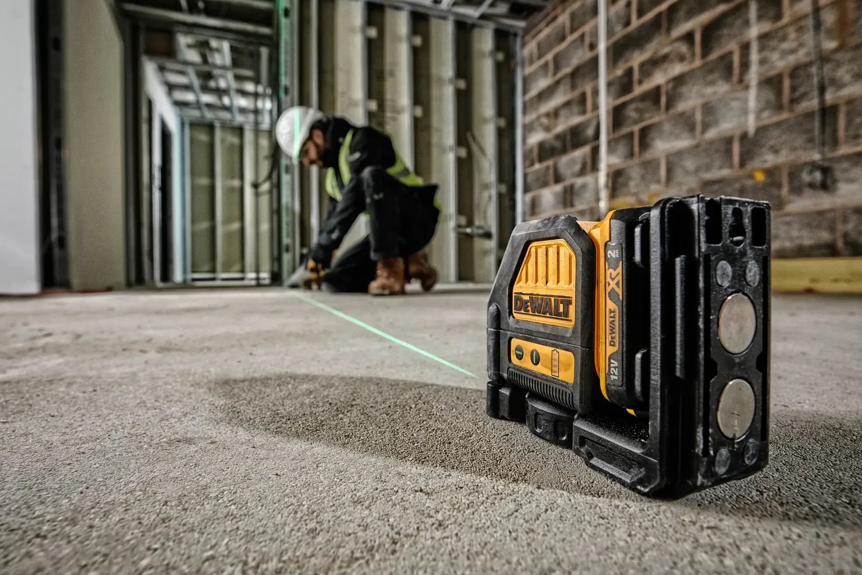 A DEWALT DCE088D1G green beam cross line laser level is placed on the floor at a construction site. In the background, a worker wearing safety equipment is using the laser line for alignment.