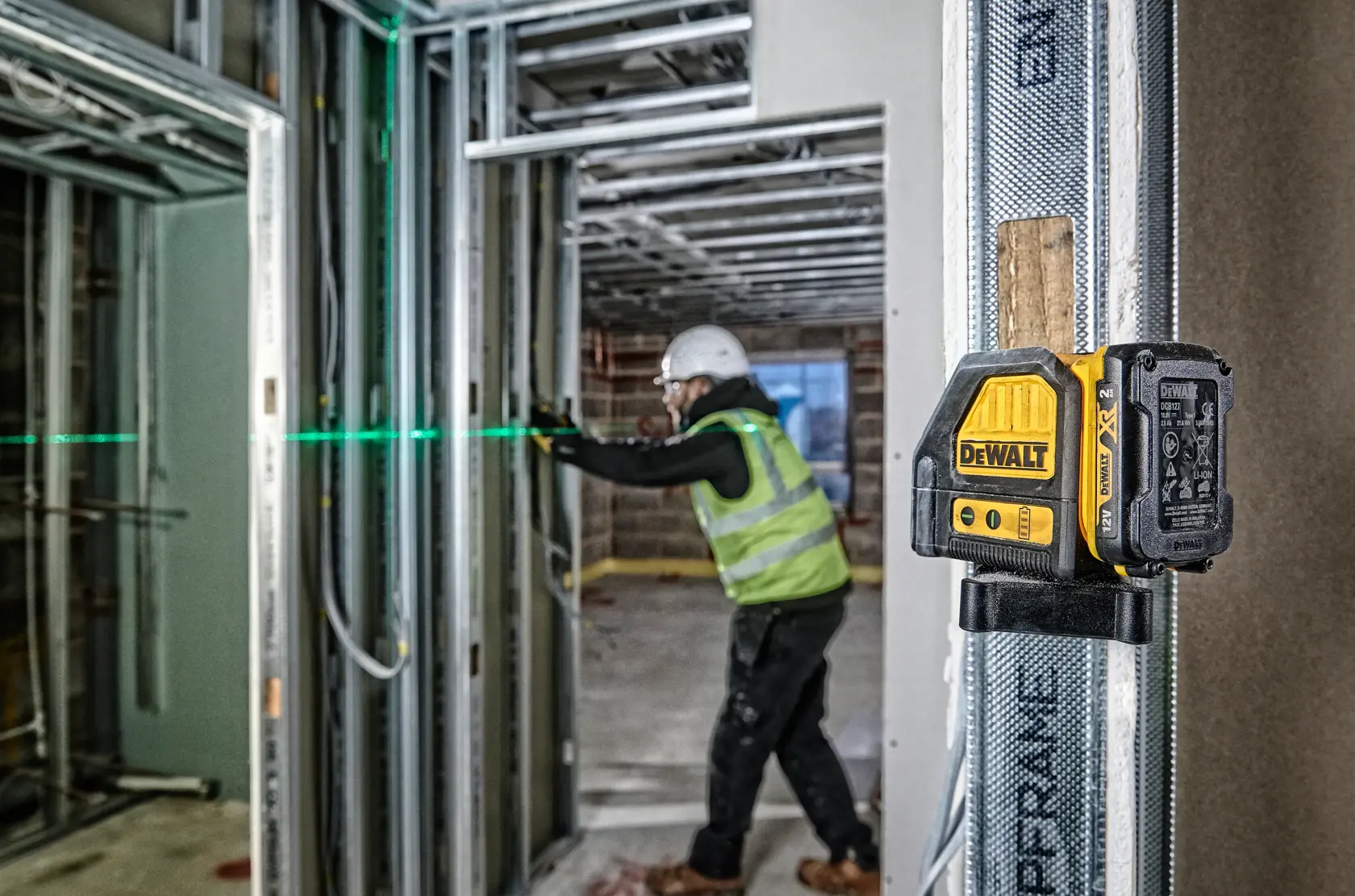 A DEWALT DCE088D1G green laser level mounted on a metal frame at a construction site, projecting a green horizontal laser line. In the background, a construction worker wearing a safety vest and helmet is aligning wall frameworks.
