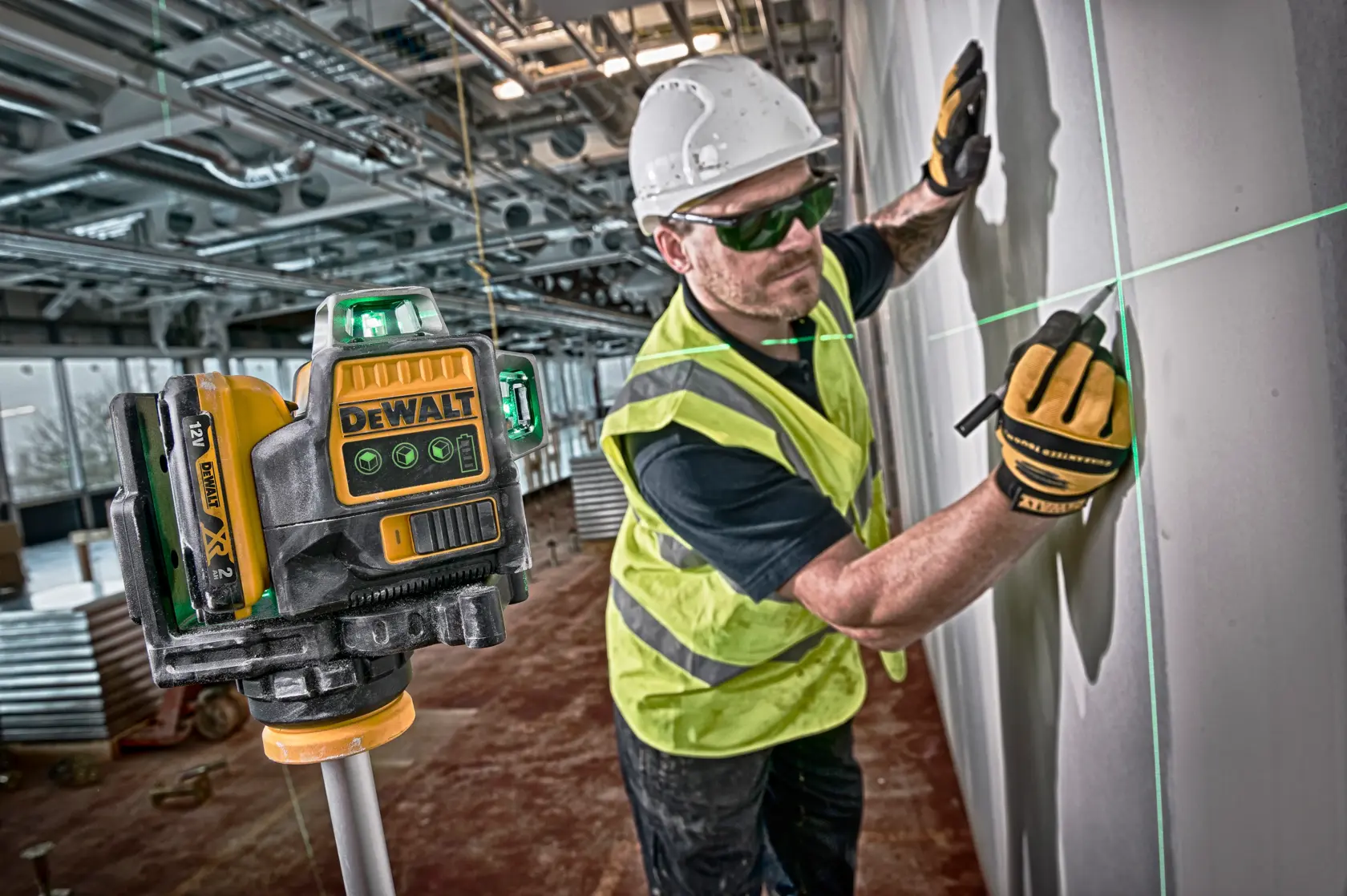 A DEWALT laser level tool is positioned in the foreground, projecting green laser lines onto a wall. In the background, a construction worker wearing a hard hat, safety vest, and gloves is marking the wall along the laser line inside an industrial setting.