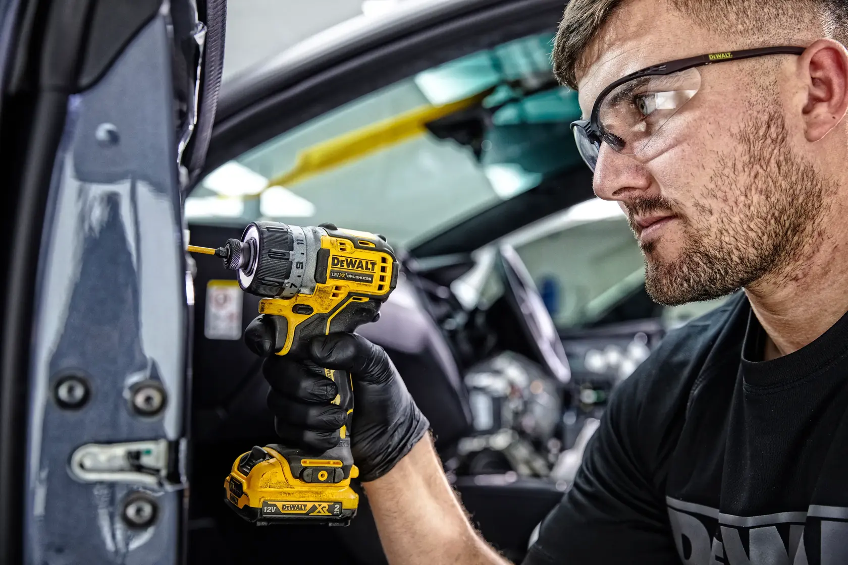 A person wearing black gloves uses a yellow DEWALT DCF601D2 cordless screwdriver to work on a part inside a vehicle. The person's face is blurred for privacy.