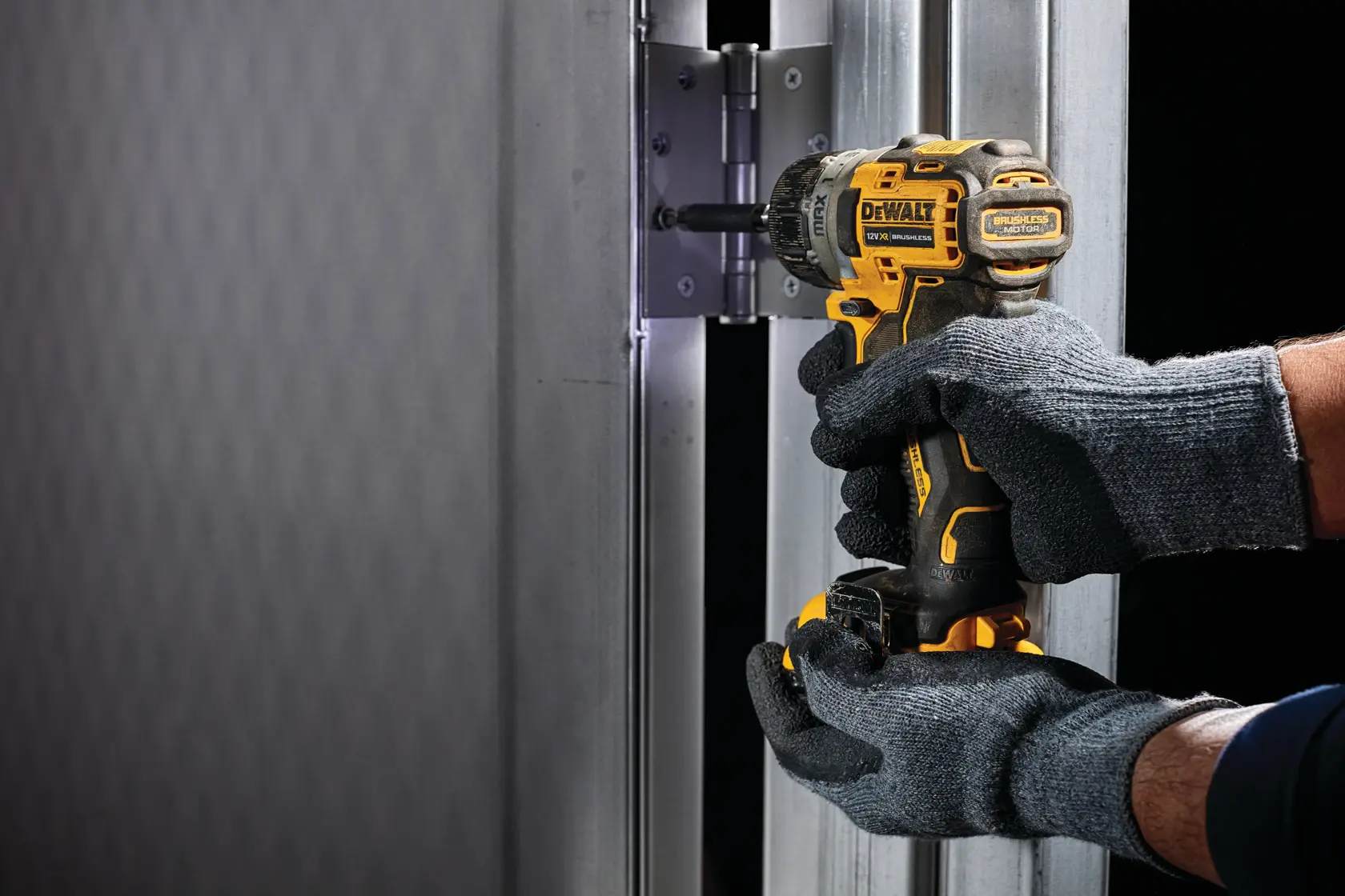 Gloved hands use a DEWALT drill to install a hinge on a metal door frame.