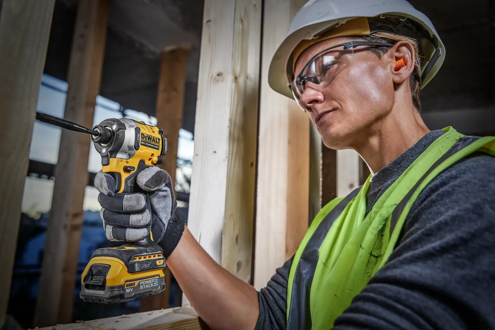 A person wearing a safety helmet, neon safety vest, and gloves uses a DEWALT DCF850E2T 18V cordless impact driver to work on